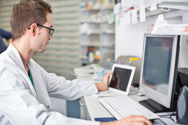 man in lab coat in front of computer terminal comparing information to data on tablet screen
